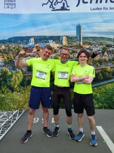 Drei jubelnde Läufer in neongelben Laufshirts mit Firmenlogo. Im Hintergrund ist ein Transparent mit der Stadt Jena zu erkennen.