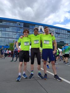 Drei lächelnde Läufer in neongelben Laufshirts draußen vor dem Ernst-Abbe-Sportfeld. Anjo hat die Arme freundschaftlich um die beiden anderen Teammitglieder gelegt